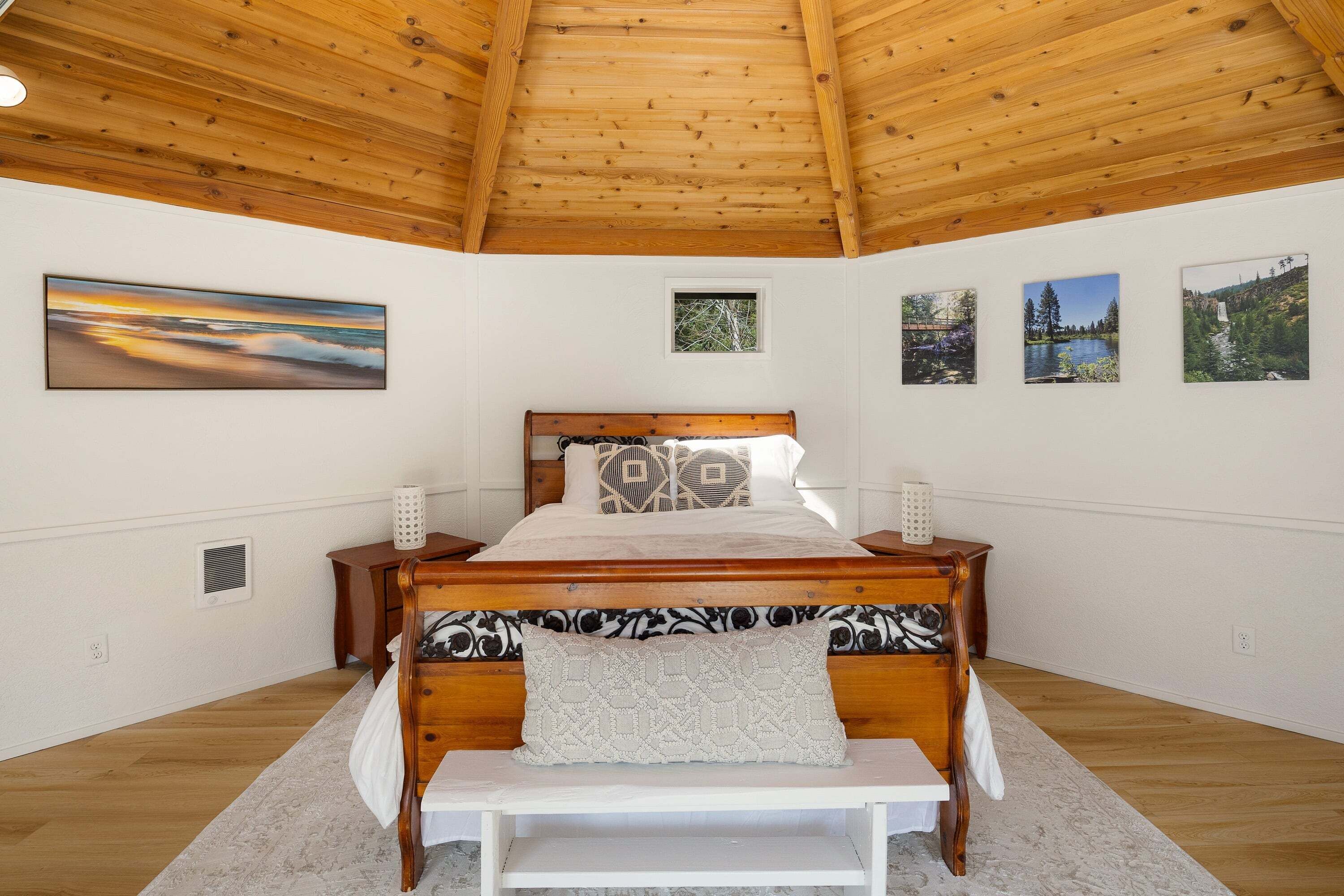 A bedroom with a wooden ceiling, a bed with patterned bedding, a bench, and three framed pictures on the wall.