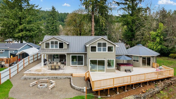 A two-story house with a deck, a patio with seating, and a fire pit. - River's Rest - Waterfront Views, Hot Tub, Grill & Fire Pit 3 Bedroom Home (Walterville)