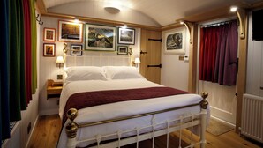 Una cama bien tendida, con cabecera y piecera blancos, una mesita de noche de madera con una lámpara, y una pared con cuadros enmarcados.