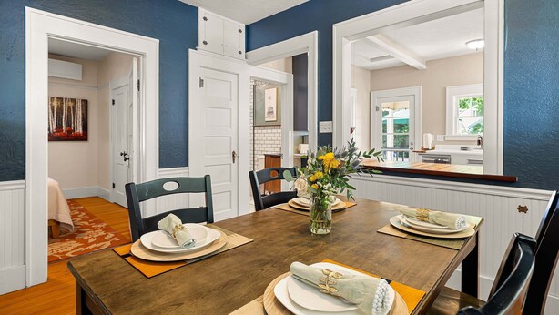 Comedor con una mesa de madera dispuesta para cuatro, armarios blancos y una cocina al fondo.