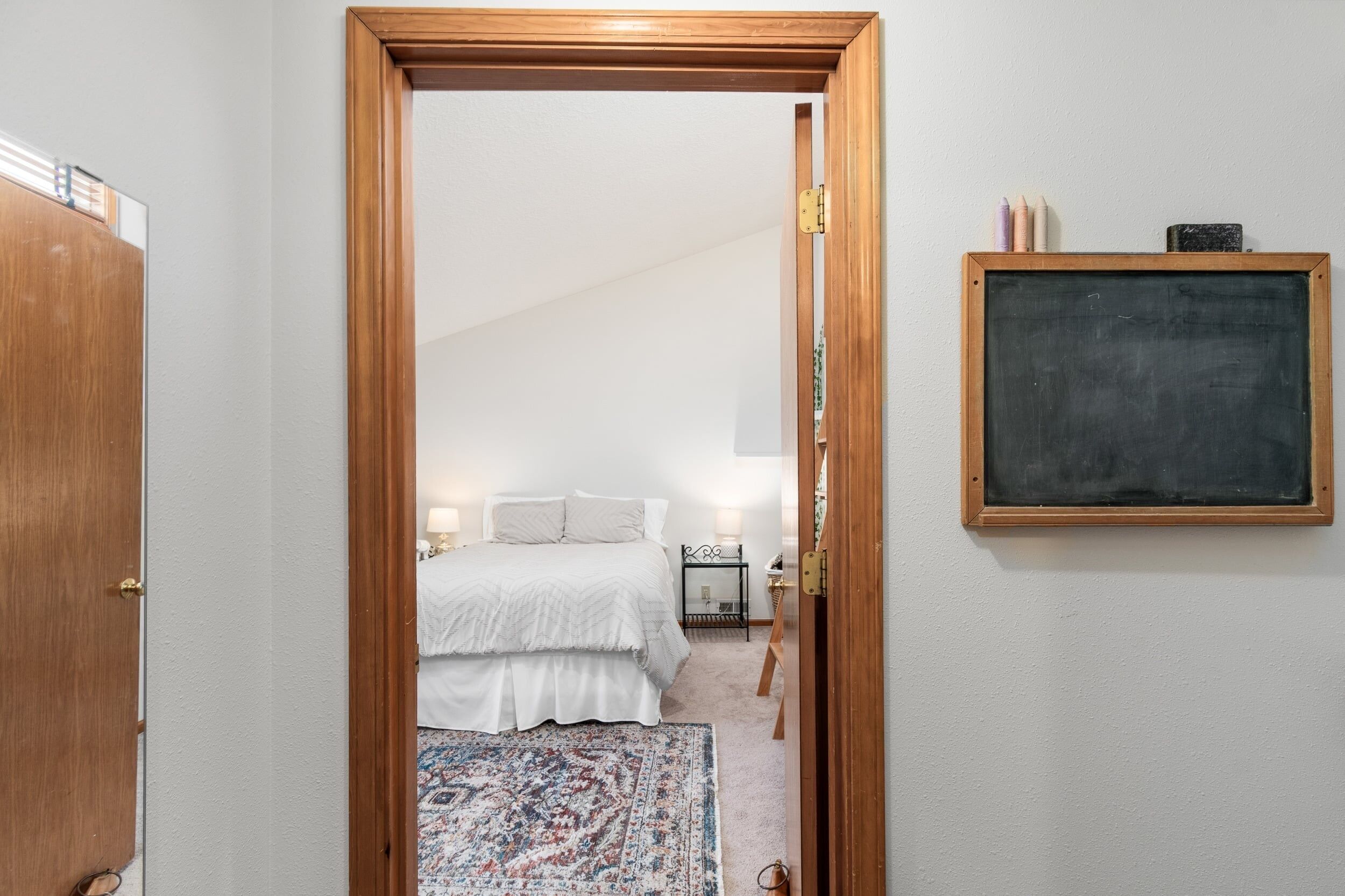Un dormitorio con cama, mesita de noche, lámpara y una pizarra en la pared.