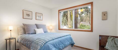 Un dormitorio con cama, lámparas de noche, una ventana que da a árboles y una mesita.