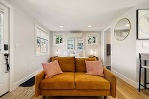 A cozy living room with a mustard-colored sofa, a bed with white bedding, and a wall-mounted mirror. - Cozy Casita - Minutes To Autzen & Downtown 1 Bedroom Home (Eugene)