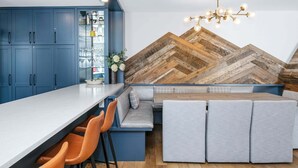 A modern kitchen with blue cabinets, a wooden herringbone-patterned wall, a bar area with stools, and a seating area with cushions. - Coldstream #18 3 Bedroom Townhouse (Vail)