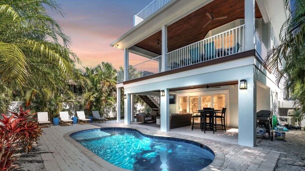Una casa de dos pisos con piscina, mobiliario exterior y un patio cubierto.
