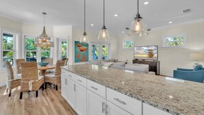 Una cocina amplia con una isla grande, comedor con sillas de mimbre y sala de estar con televisión y sofá azul.