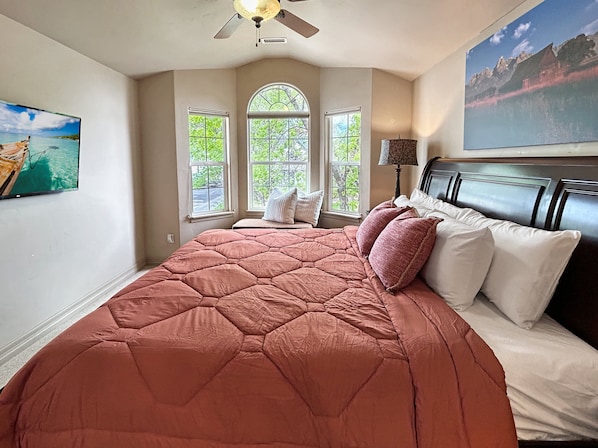 A bedroom with a large bed, a ceiling fan, a window with a view of greenery, and a painting on the wall. - Victorian Chateau Retreat 3 Bedroom Townhouse (Grand Junction)