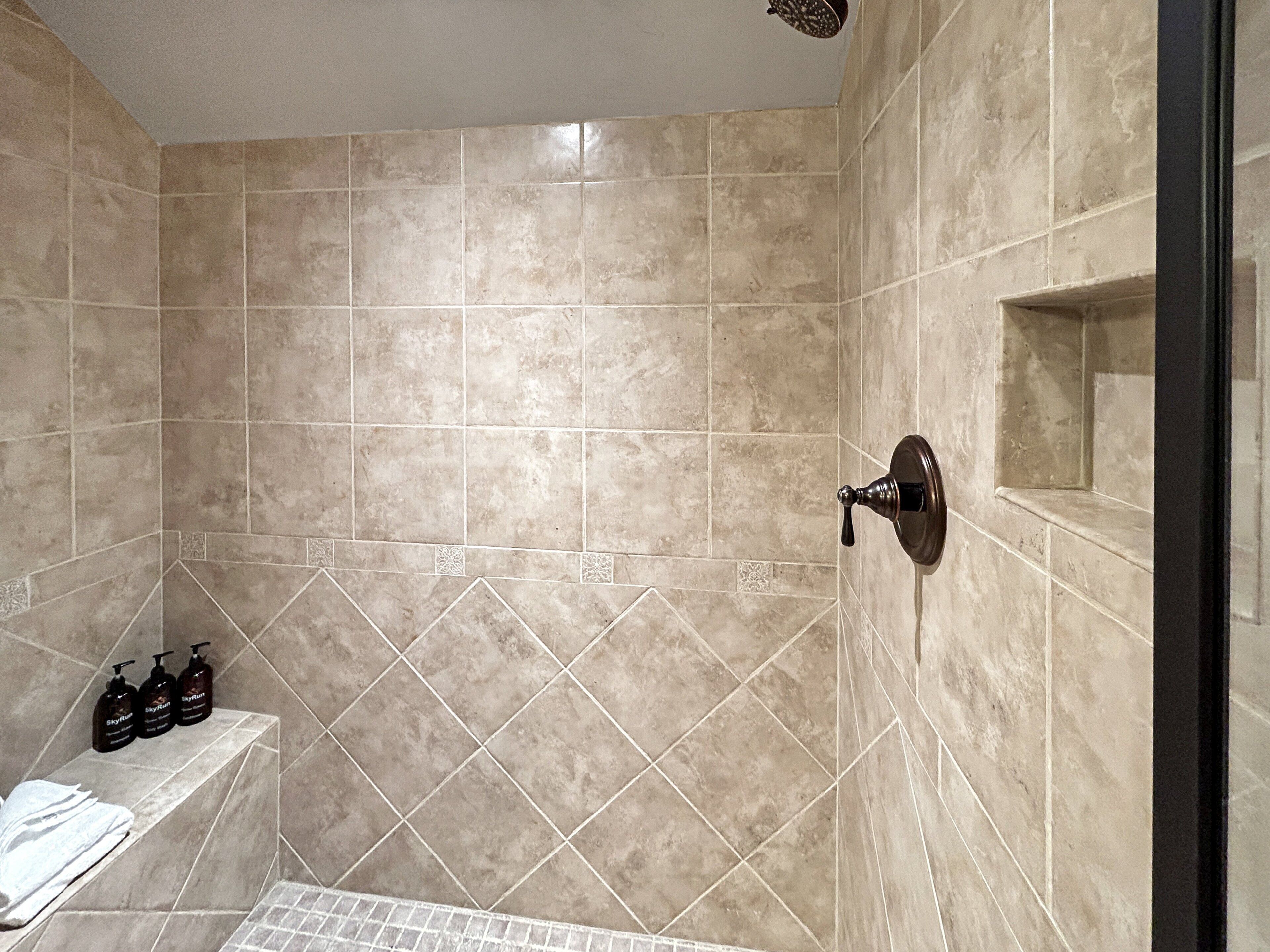 A bathroom with beige tiles, a shower control, and toiletries on a shelf.