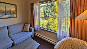 A living room with a sofa, a chair, a window with curtains, and a painting on the wall. - Cahilty #433 Hotel Room (Sun Peaks)