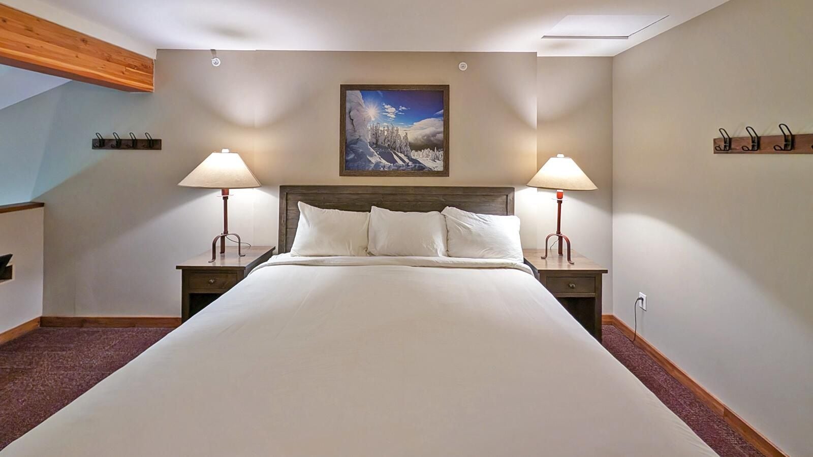 A hotel room with a large bed, two bedside tables with lamps, a painting on the wall, and a wooden ceiling beam.