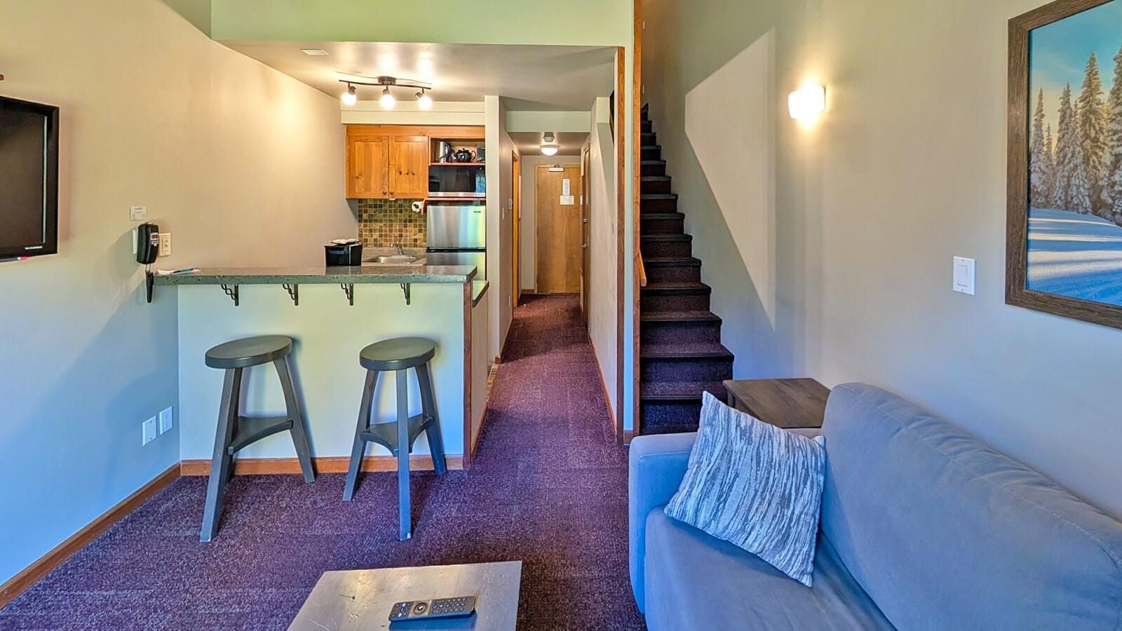 A compact living space with a kitchen, a dining area with stools, a staircase, and a sofa with a cushion.