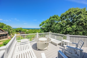 A deck with a round table and chairs, surrounded by white railings, overlooking a green landscape and the sea.