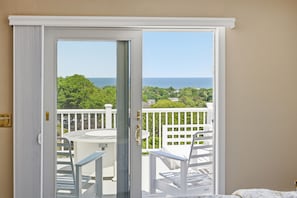 A room with a sliding glass door leading to a balcony with a view of the ocean and greenery.