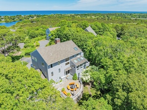 A two-story house with a large deck, surrounded by dense greenery and a view of the ocean.