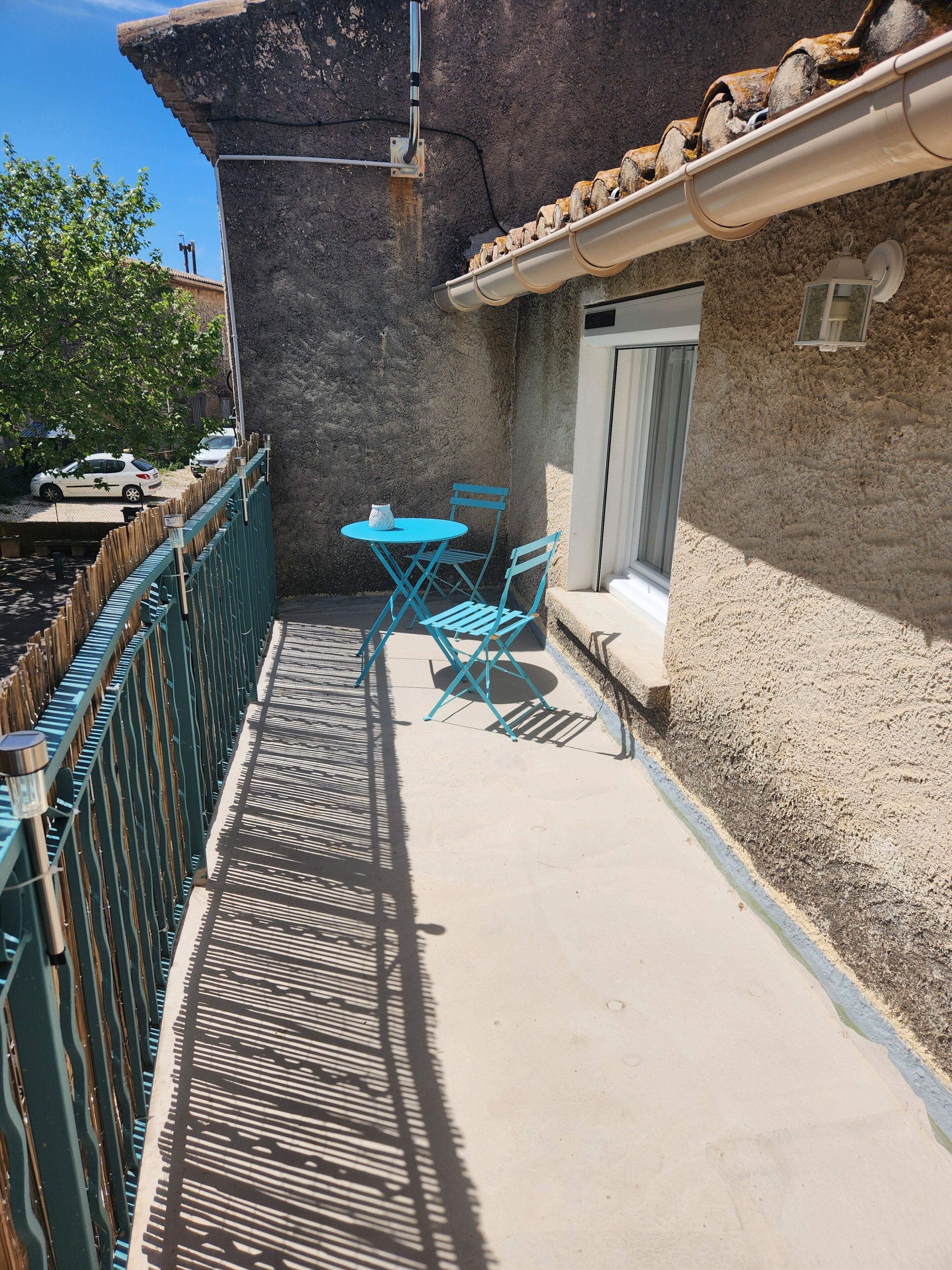 A narrow outdoor patio with a table and chairs, a metal fence, and a building with a textured wall and a window.