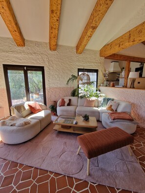 Ein gemütliches Wohnzimmer mit einer Couch, Hocker und Couchtisch, ein Küchenbereich mit Herd und Schränken sowie große Fenster mit Blick auf Grünflächen.