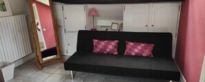 Ein Zimmer mit einer schwarzen Couch, zwei Kissen in Rosa mit Punkten, einem Holzstuhl, einem weißen Heizkörper, einer pinkfarbenen Lampe und einem weißen Kleiderschrank mit roten Griffen.