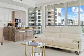 A modern living room with a sofa, a coffee table, and a kitchen area with a microwave and bar stools. - Luxury 2BR 2.5BA Amrit Condo with ocean view (Riviera Beach)