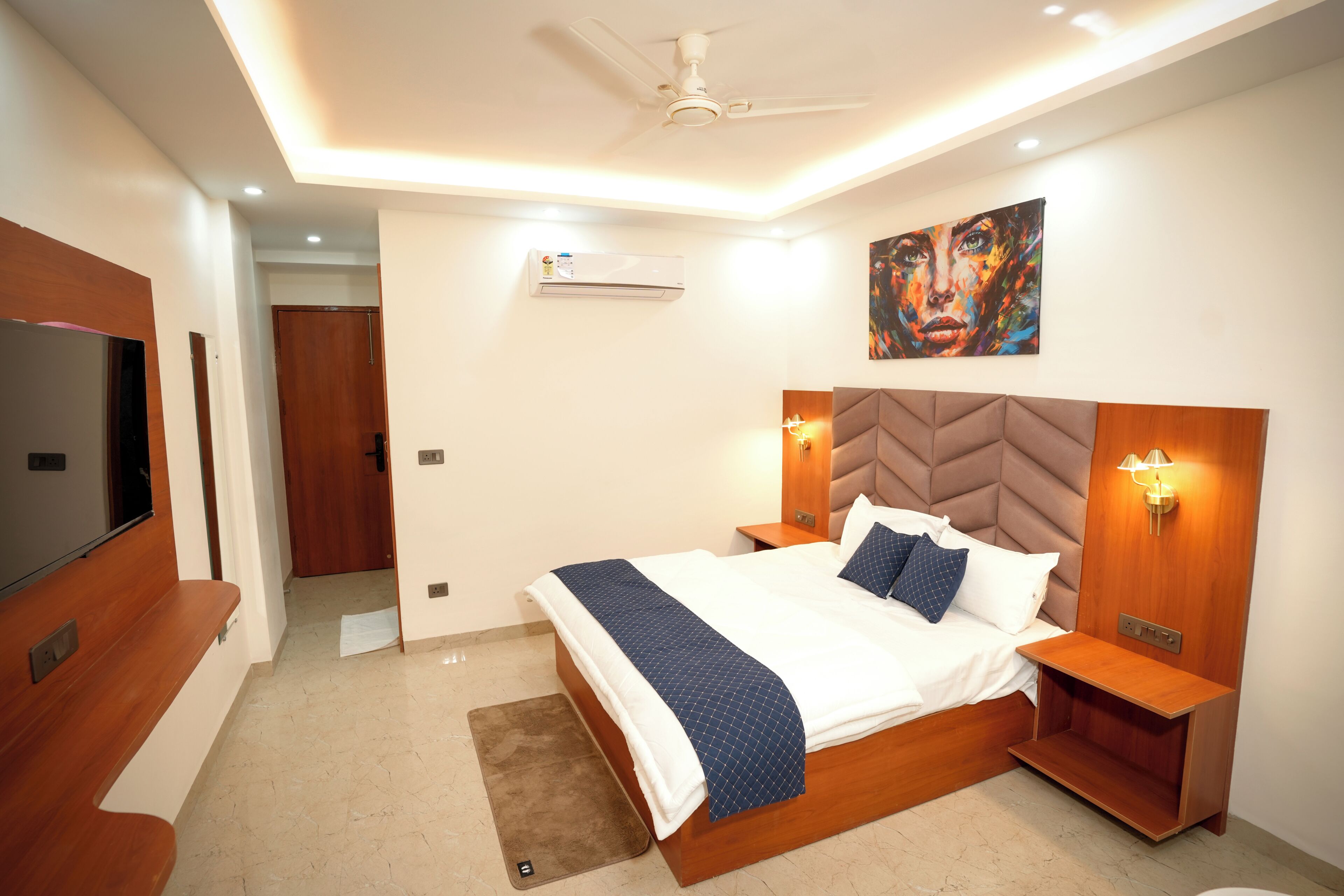 A modern bedroom with a large bed, bedside tables, a ceiling fan, an air conditioning unit, and a painting on the wall.
