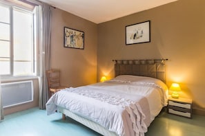 A bedroom with a bed, two bedside lamps, a chair, a radiator, and two framed pictures on the wall.