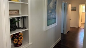 A hallway with white walls, a framed painting of a cloudy sky, a shelf with decorative items, and a wooden floor. - Marvelous 3-bedroom, newly renovated house with AC in charming Tyler (Tyler)