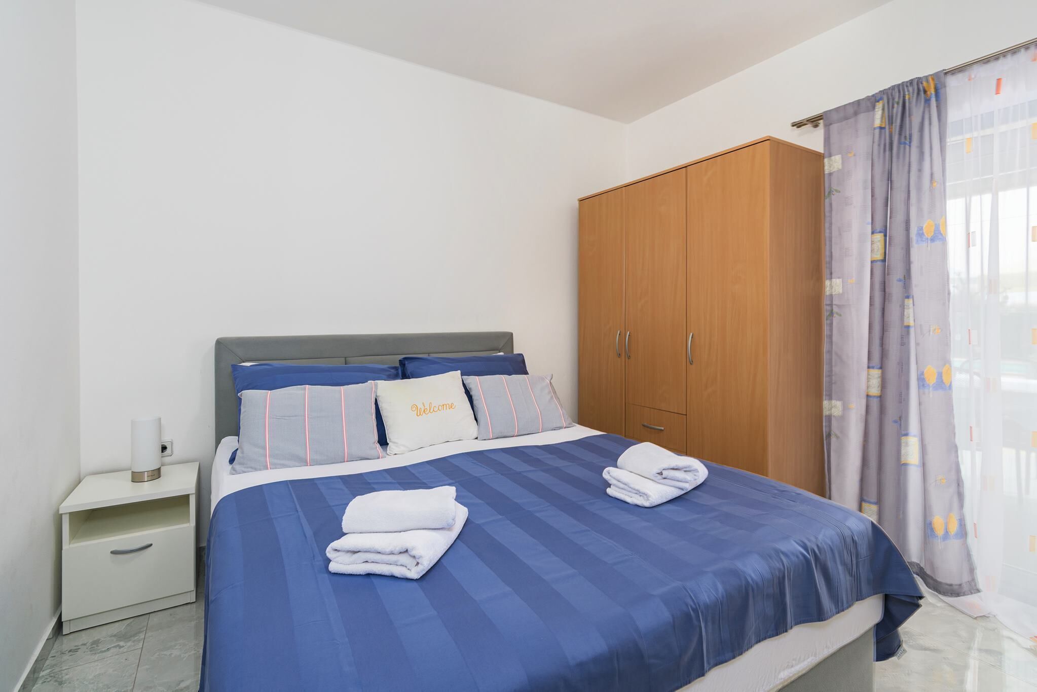 Un lit bien fait, avec une literie bleue et un coussin orné de la mention « Bienvenue », une armoire en bois, une table de chevet avec une lampe et une fenêtre agrémentée de rideaux à motifs.
