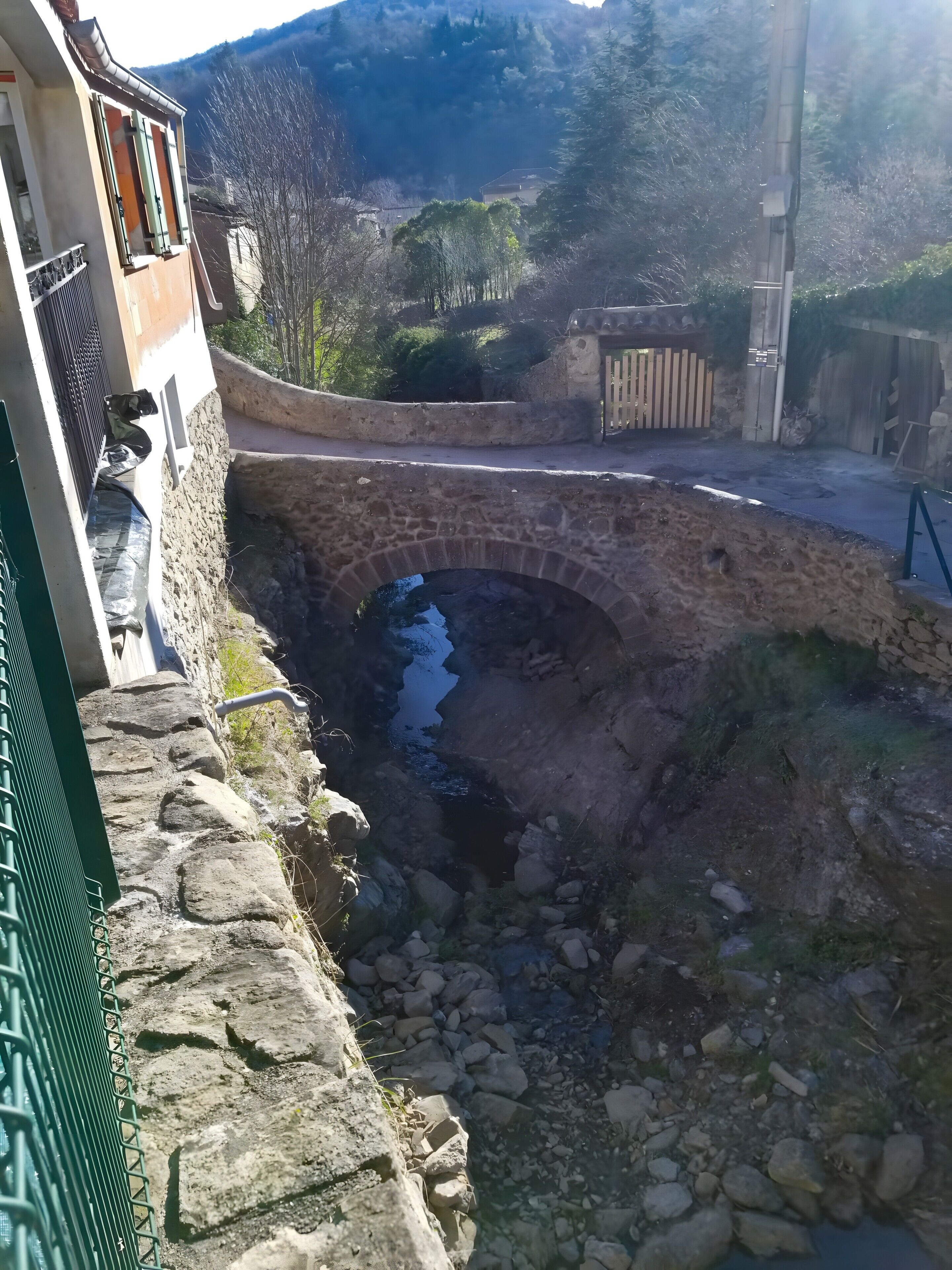Un puente de piedra sobre un pequeño arroyo, un muro de piedra y un edificio con balcón.