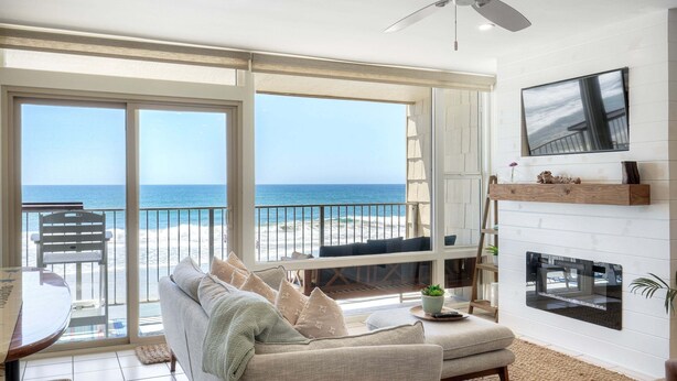 Un salón con un ventanal grande que da a una playa, un ventilador de techo y una chimenea.