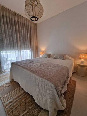 Ein Schlafzimmer mit einem großen Bett, zwei Nachttischlampen, einem fensterbelaubten Fenster und einer Hängelampe.