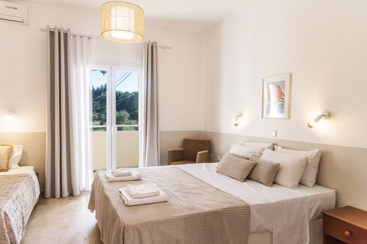 Una habitación de hotel con cama, una silla, un escritorio, una ventana con cortinas y un cuadro colgado en la pared.