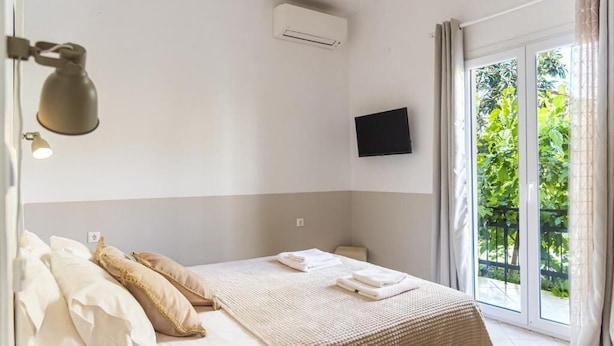 A bedroom with a bed, pillows, a flat-screen TV, an air conditioning unit, and a view of greenery through a sliding glass door. - BOHOTEL Eleonas Anex Apartments (Corfu)