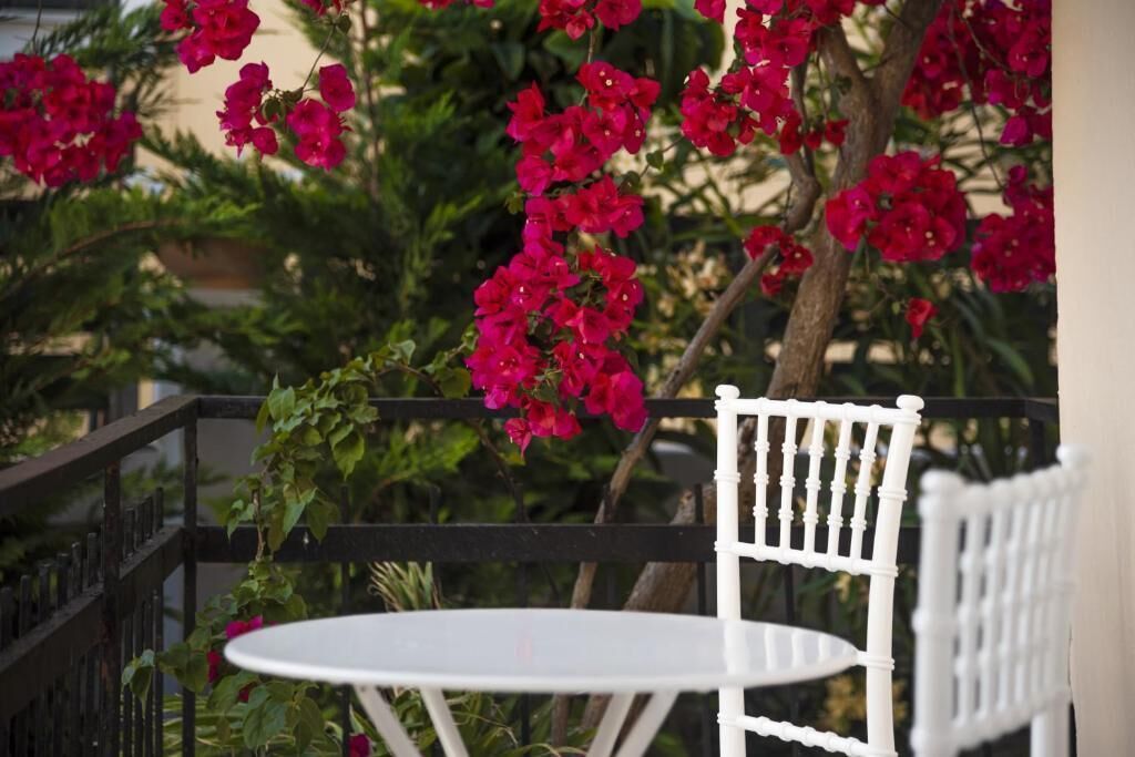 Une table et des chaises blanches sont disposées sur un balcon, avec des fleurs rouges en arrière-plan.