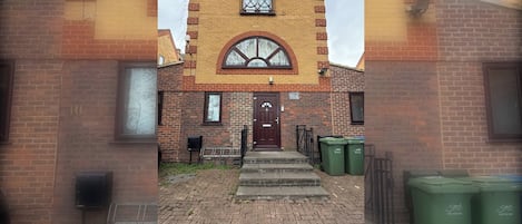A brick building with a central arched window and a door, surrounded by a paved area with steps leading to the entrance.