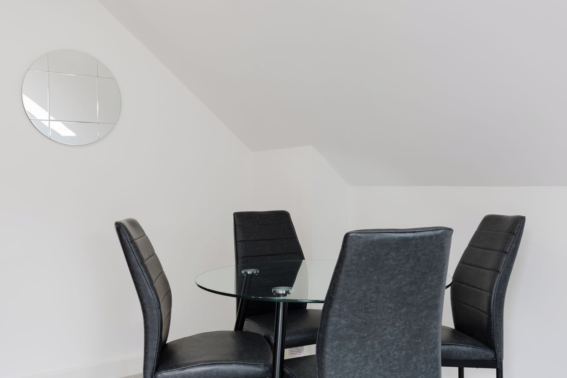 A dining area with a round table and four chairs, a wall-mounted mirror, and a sloped ceiling.