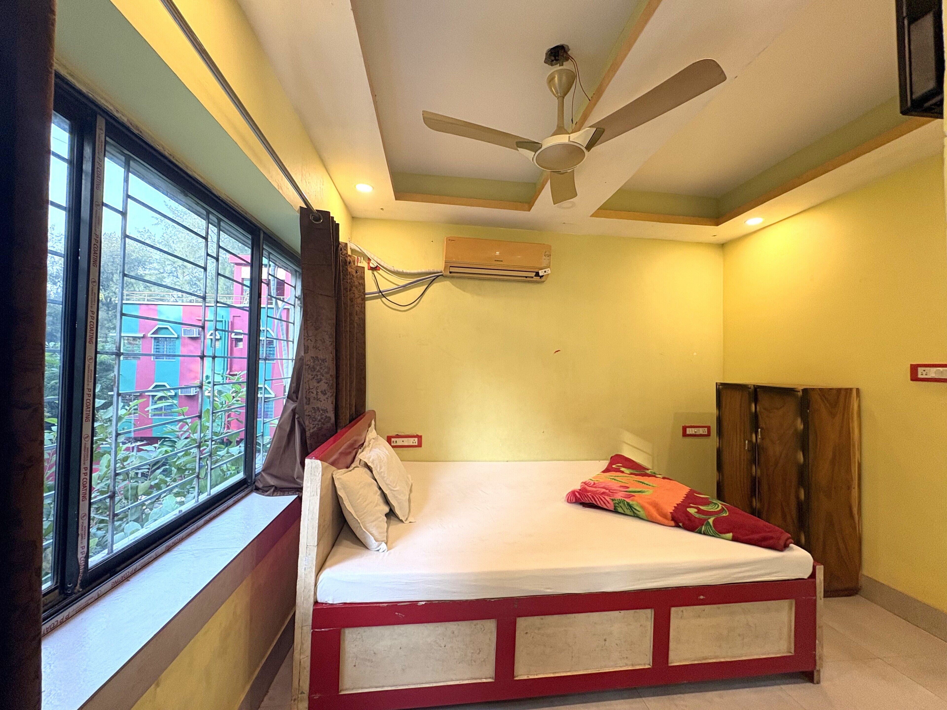 Una habitación con cama, aire acondicionado, ventilador de techo y ventana con vistas a edificios.