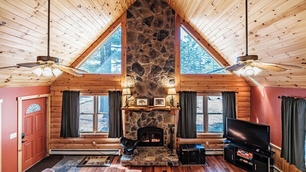 A log cabin interior with a stone fireplace, wooden walls, a television, and a ceiling fan. - Riverwood Chalet- Near Sunday River (Newry)