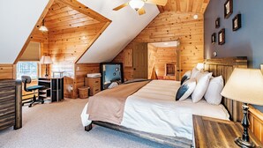 A room with a sloped ceiling, wooden walls, a bed with pillows, a desk with a chair, and a ceiling fan. - Riverwood Chalet- Near Sunday River (Newry)