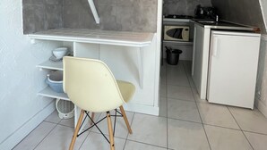 Cocina compacta con refrigerador blanco, estufa y lavabo. Pequeño comedor con una silla y una estantería.
