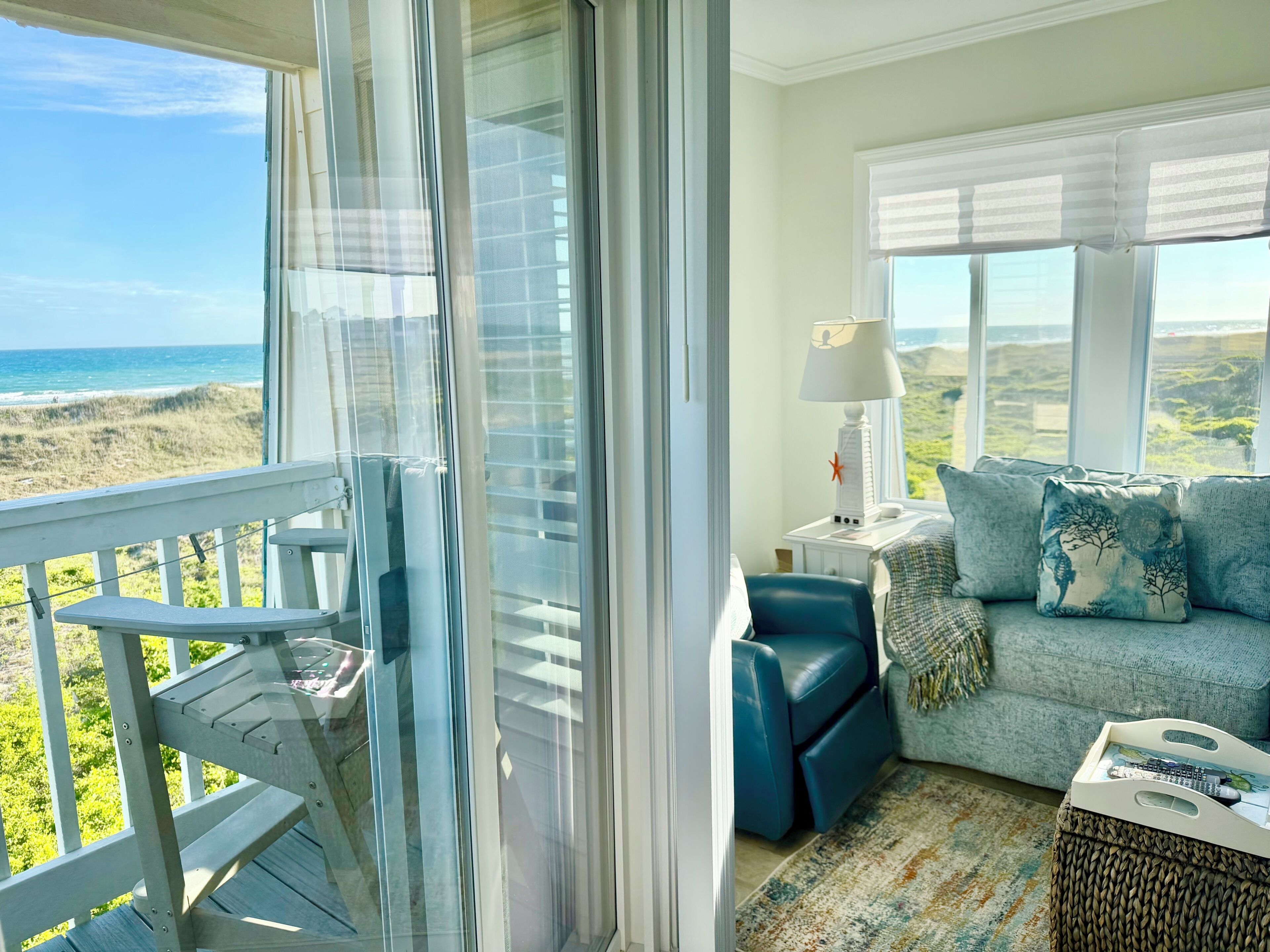 Ein Balkon mit Blick auf den Strand, eine Couch mit Kissen, eine Lampe und ein gewebter Hocker.