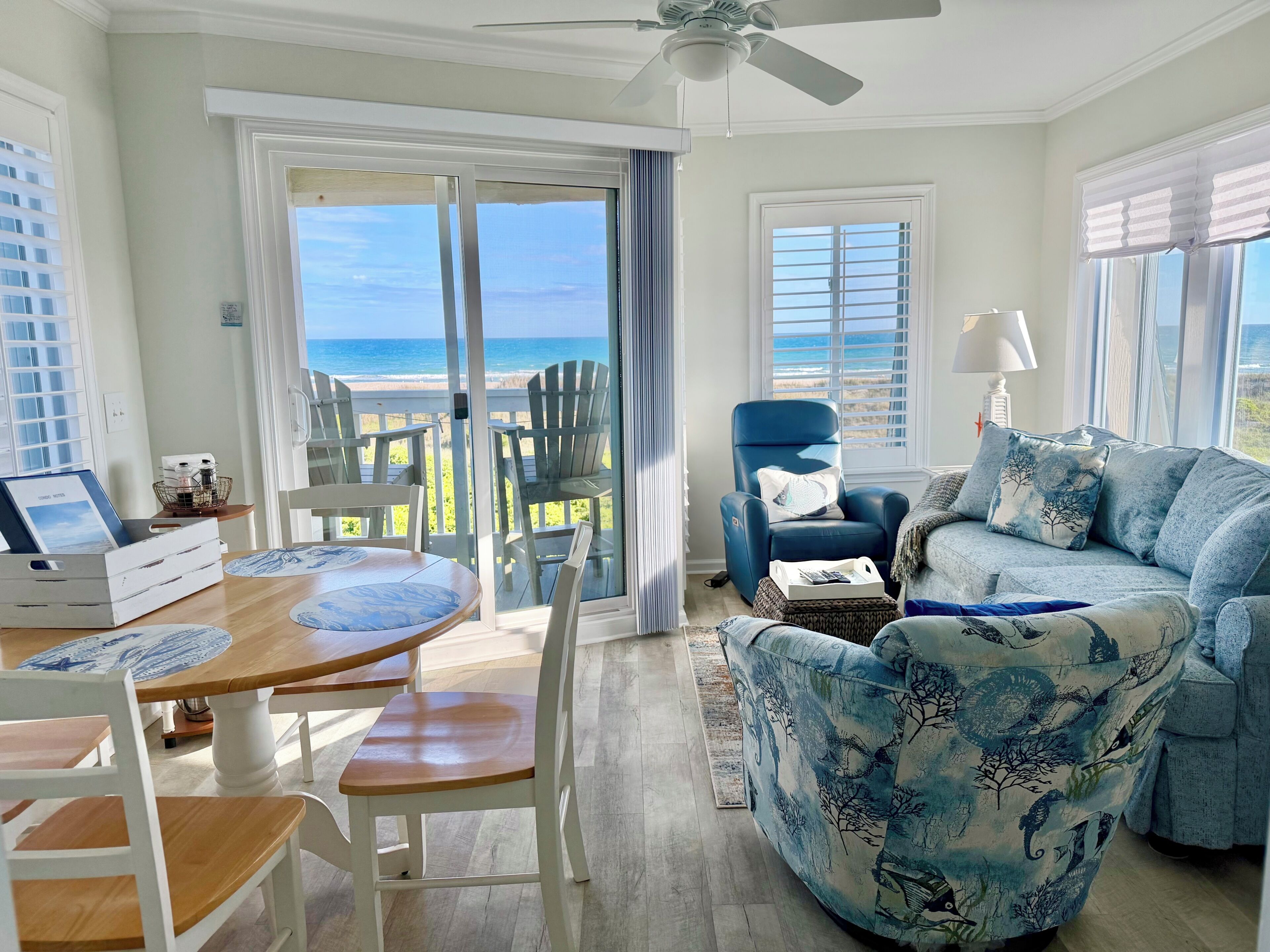 Ein Zimmer mit Blick auf den Ozean, einem Deckenventilator, einem blauen Sessel, einem Holzesstisch und einem Sofa mit blau-weißem Muster.