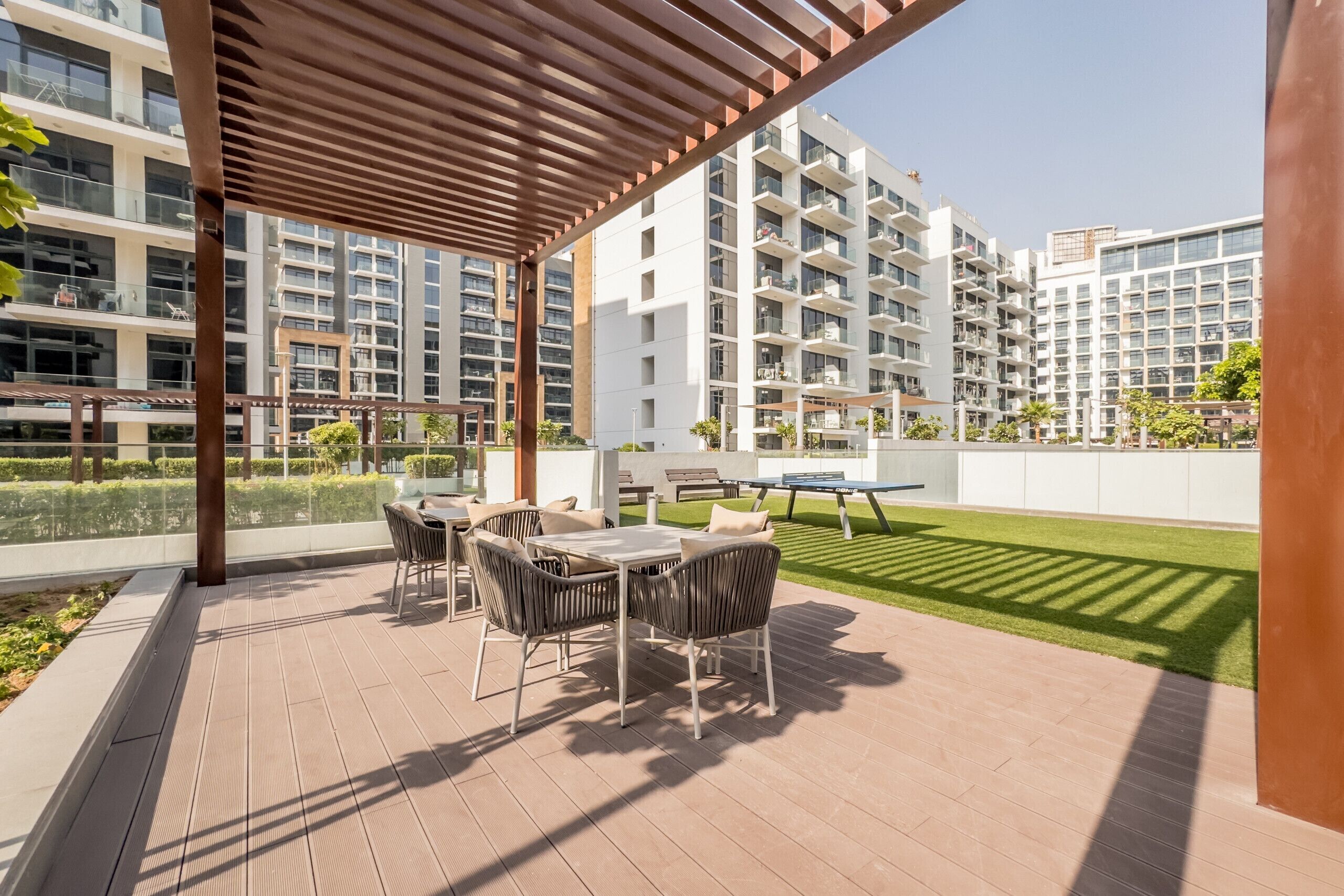 A covered outdoor area with a table and chairs, a lawn, and high-rise buildings in the background.