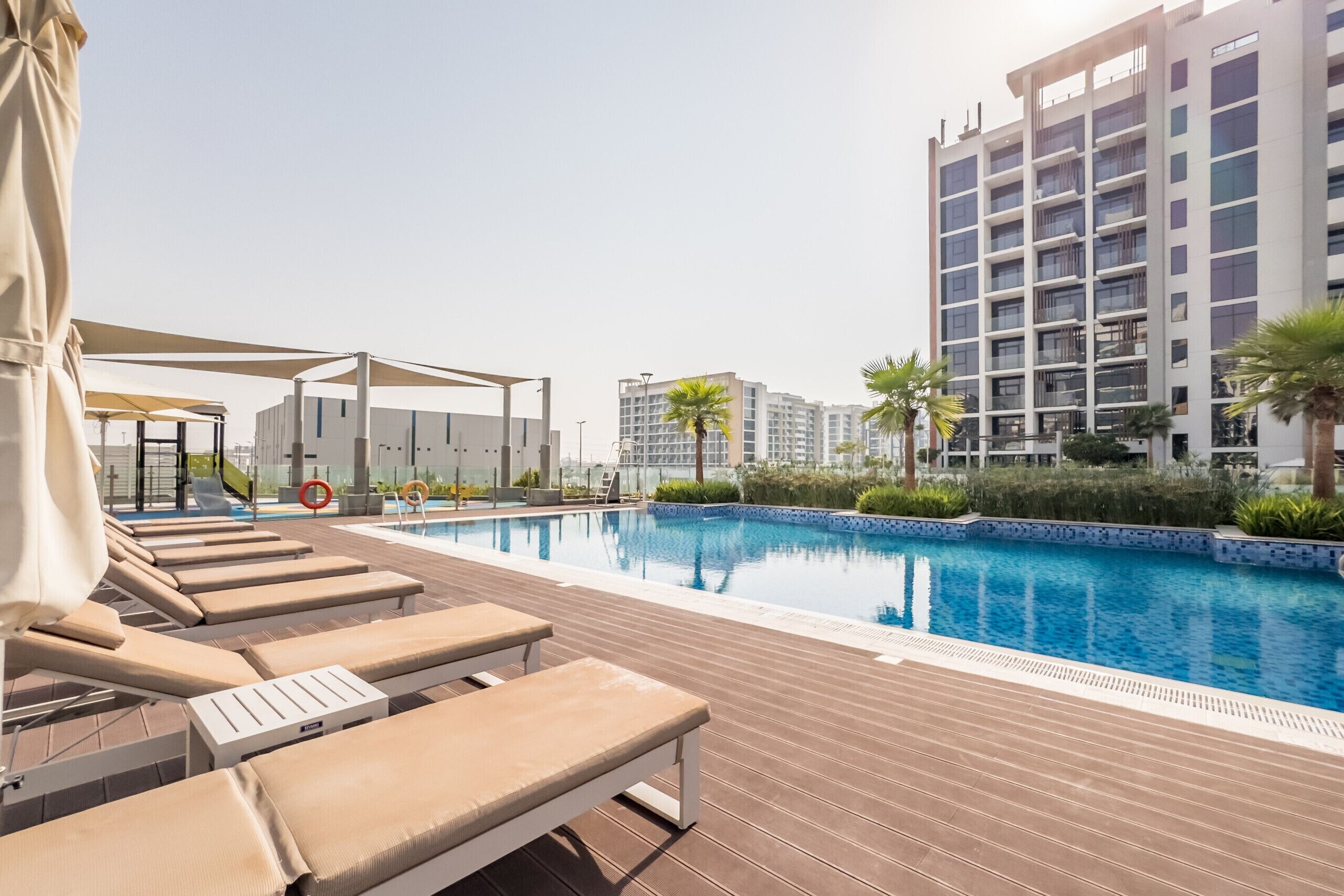 A swimming pool with lounge chairs, a covered seating area, and modern buildings in the background.