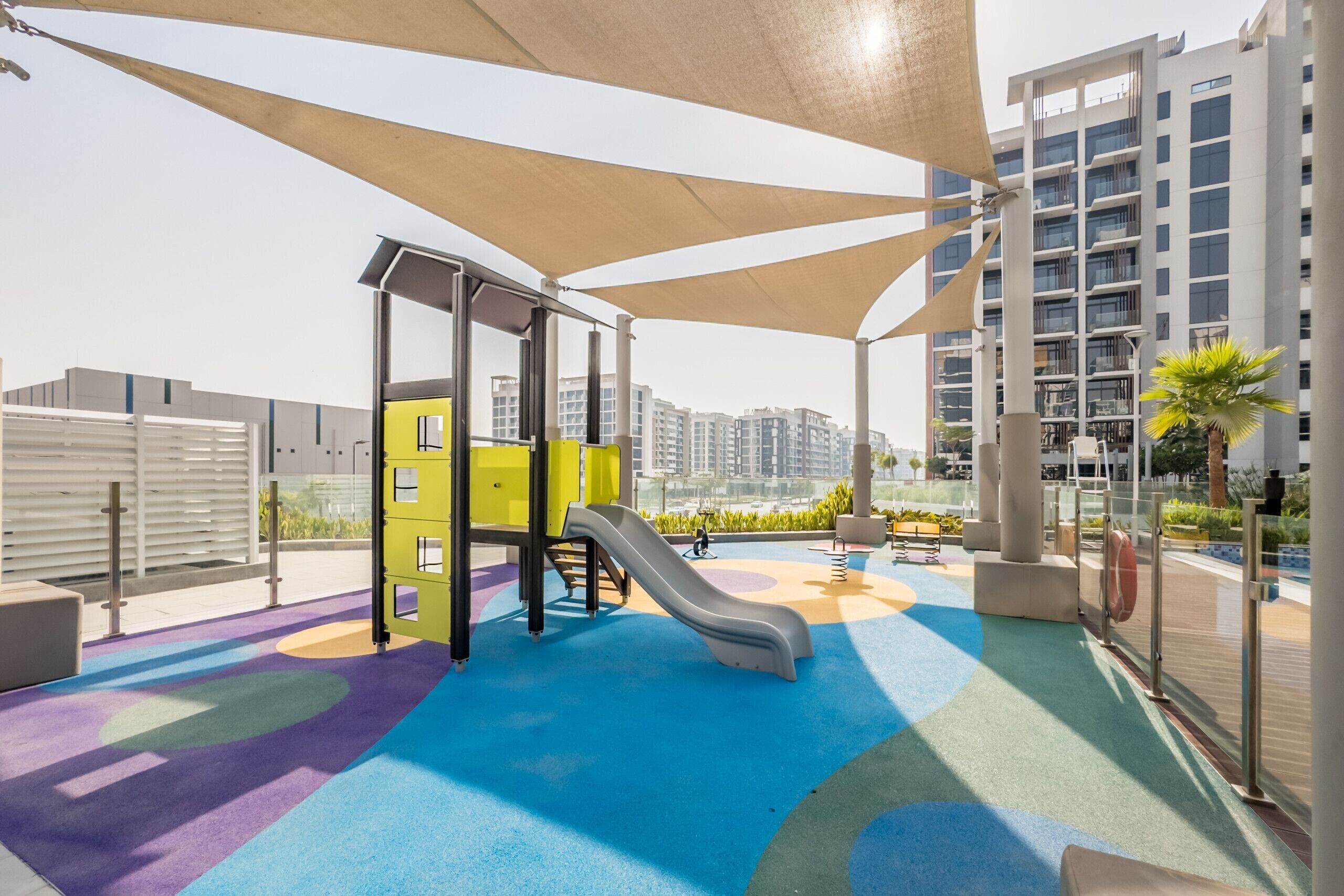 A children's play area with a slide and climbing structure, surrounded by a colorful floor and shaded by large canopies.