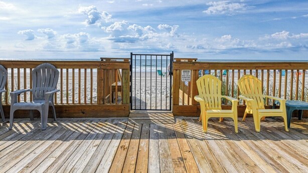 Een houten terras met stoelen en een tafel, met uitzicht op een strand en heldere lucht.