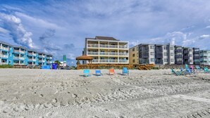 Een hotel aan het strand met meerdere balkons, strandstoelen en een zandstrand.