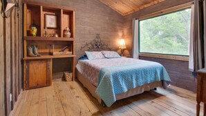 Un dormitorio de cabaña de madera con una cama, estanterías con botas, una lámpara y una ventana con vistas a árboles.