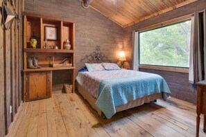 Ein Schlafzimmer einer Holzhütte mit einem Bett, Regalen mit Stiefeln, einer Lampe und einem Fenster mit Blick auf Bäume.