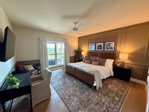 Ein Schlafzimmer mit einem Bett, einem Schreibtisch, einem Sessel, einem Deckenventilator und einem großen Fenster mit Vorhängen.