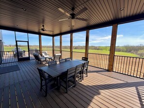 Eine überdachte Terrasse mit Tisch und Stühlen, einem Deckenventilator und Blick auf ein grünes Feld.
