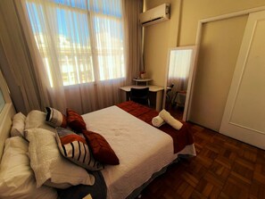 Quarto de hotel com uma cama, uma mesinha, uma cadeira e um espelho.